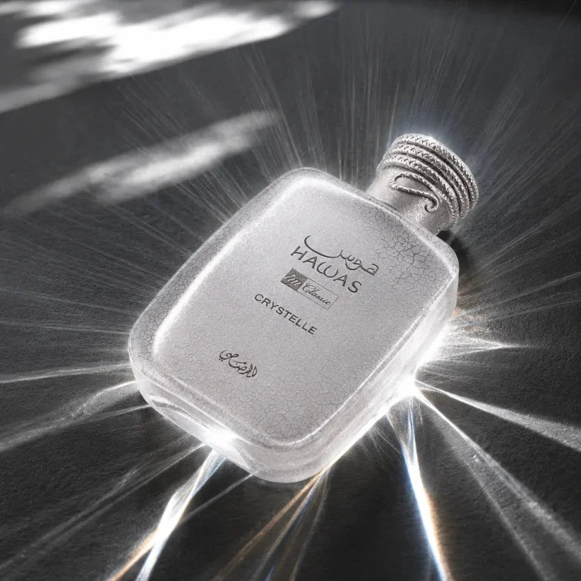 A silver-toned rectangular perfume bottle with a textured cap, featuring the brand name 'HALAS' and 'CRYSTALLE' printed on the front, set against a dark reflective background with dramatic light rays.