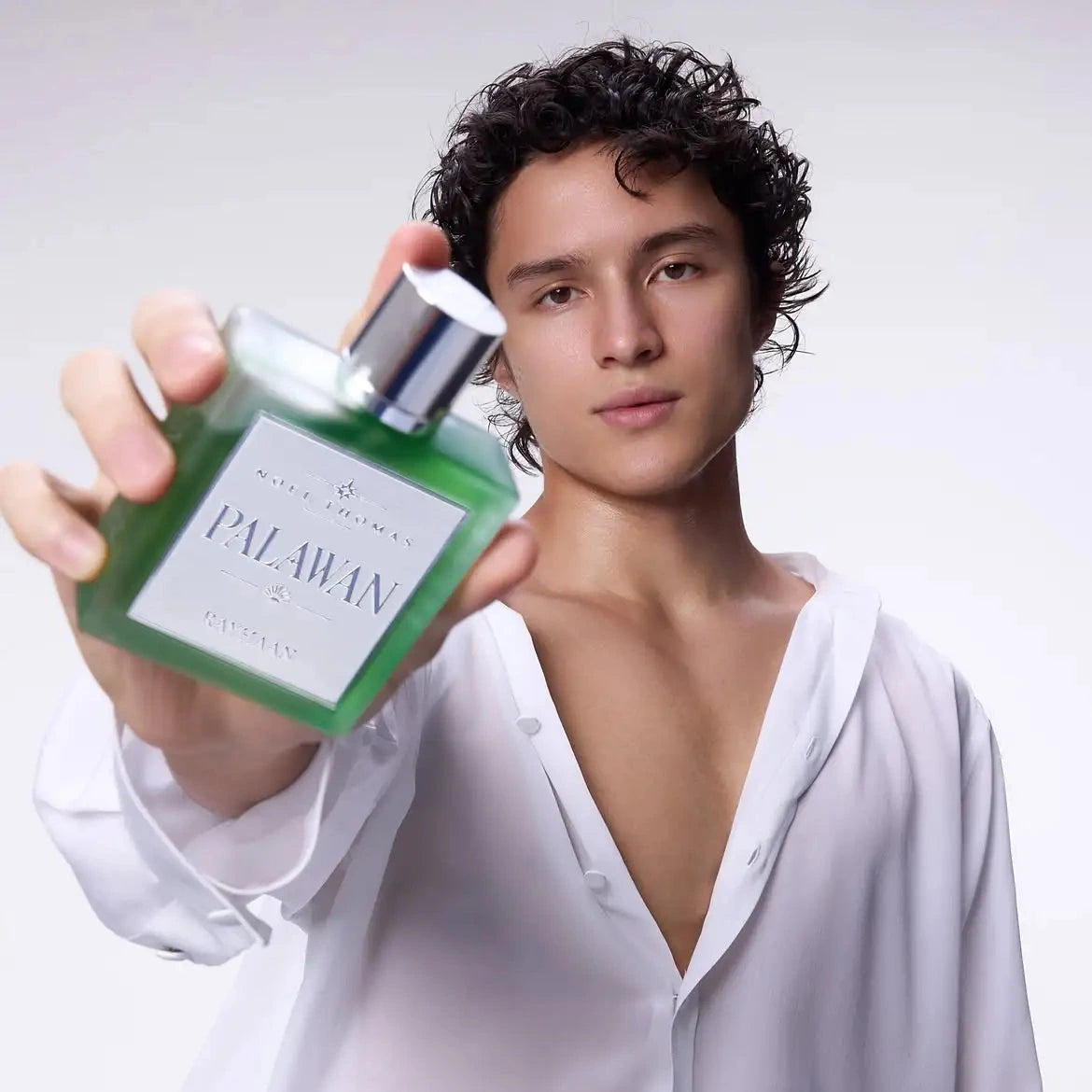 A man in a white shirt holds up a green rectangular perfume bottle labeled 'PALAWAN' against a light gray studio background.